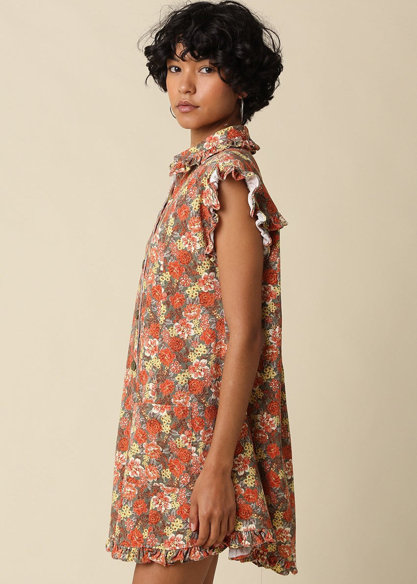 A woman wearing a sleeveless, high-neck mini dress with a dense floral print in red, orange, and yellow on a beige background, photographed against a solid light beige studio backdrop.
