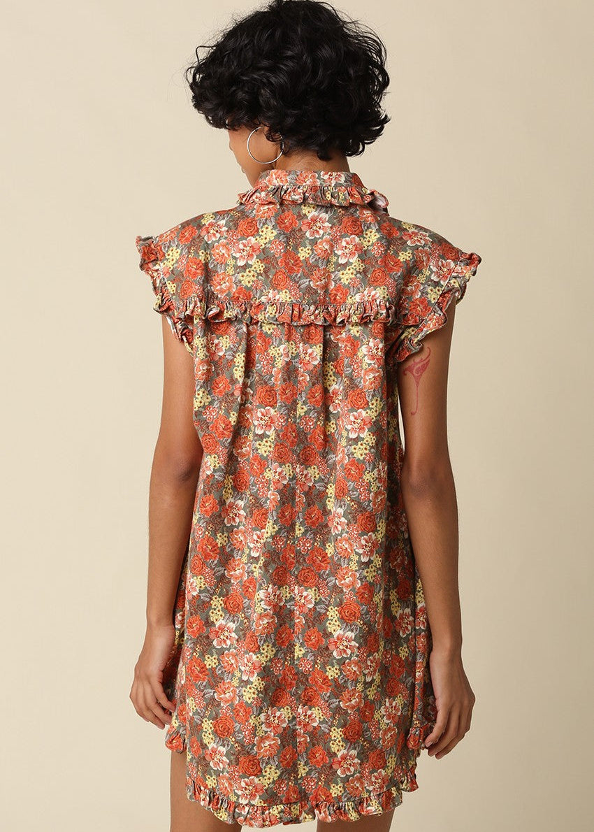 A woman shown from the back wearing a sleeveless ruffled dress with an all-over floral print in rust red, orange, cream, and olive tones against a plain beige studio background.