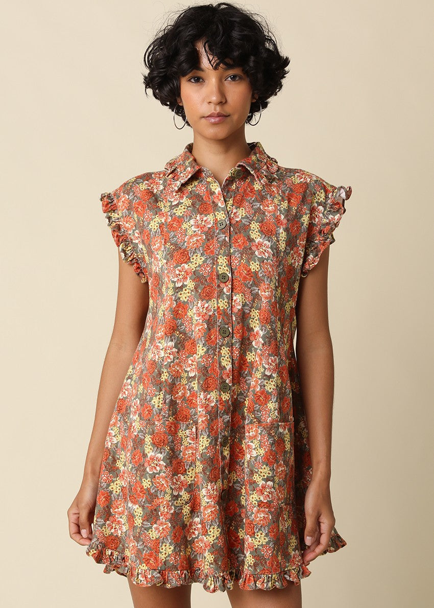 A woman modeling a short-sleeve button-up shirt dress with a dense floral print in orange, red, yellow, and green on a beige base, set against a solid light beige background.