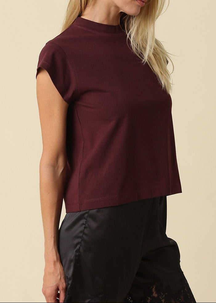 A woman wearing a short-sleeve crewneck top in deep burgundy, paired with black pants, photographed against a solid light beige background.