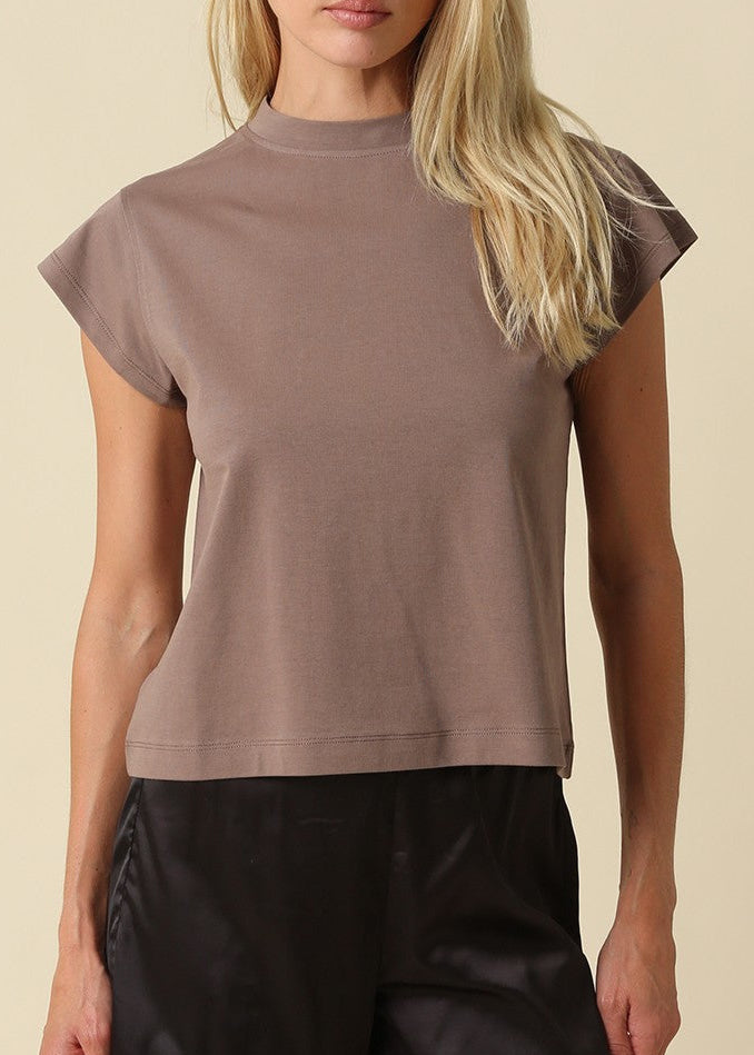 A blonde model wearing a taupe-brown short-sleeve mock neck crop top and black pants, standing against a solid beige background.