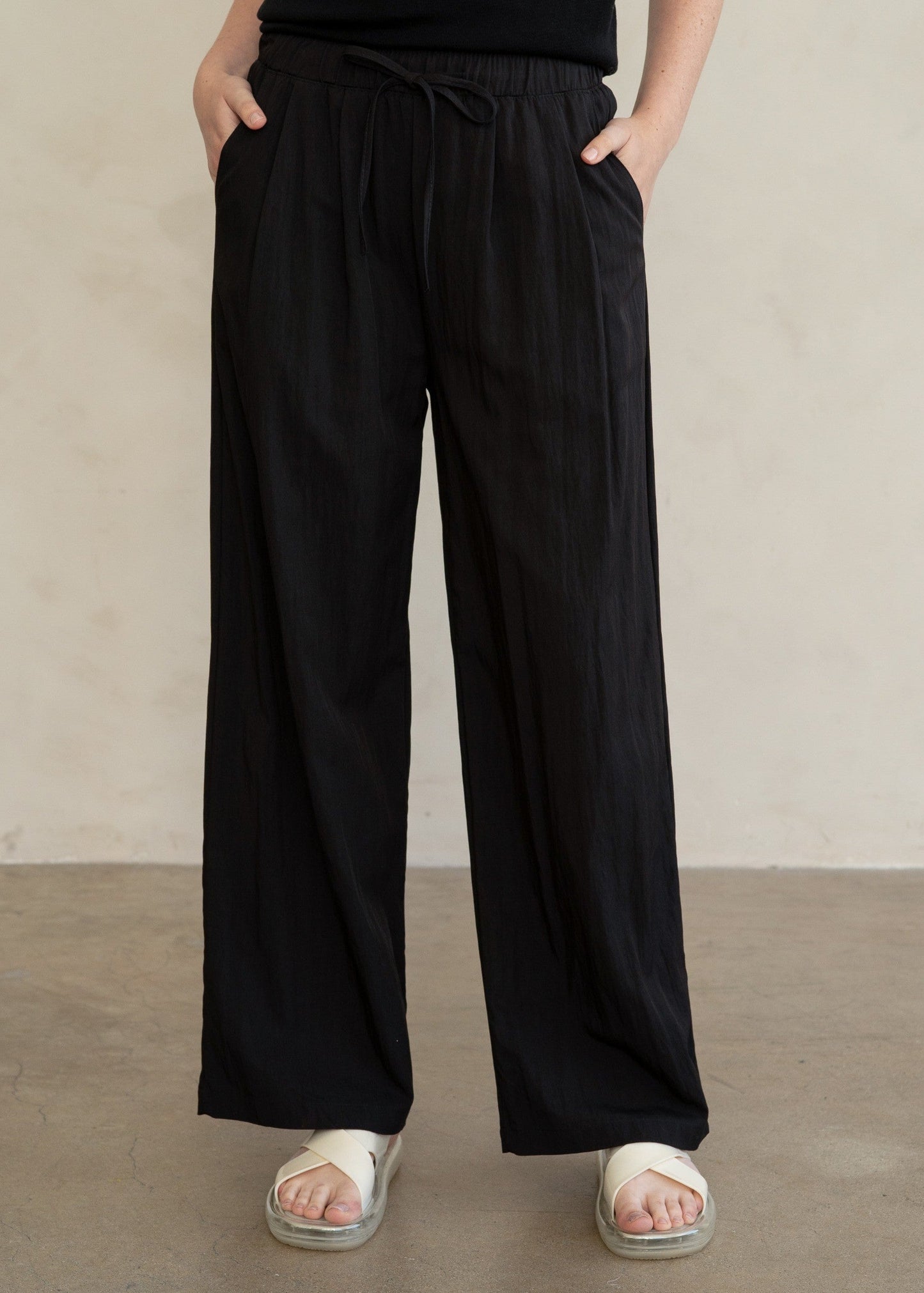A person wearing loose black wide-leg pants with an elastic drawstring waist, paired with a black sleeveless top and white slide sandals, standing against a plain light beige wall.