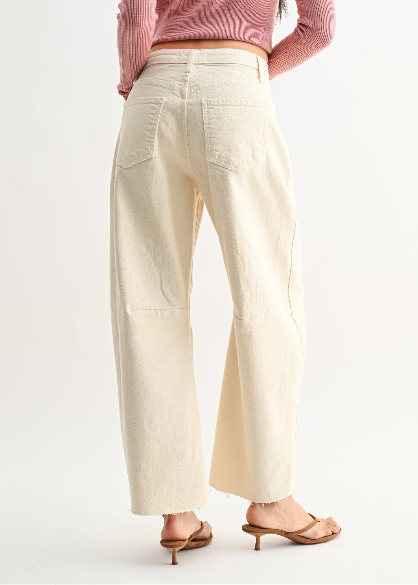 Cream-colored barrel jeans with high waist, visible back pockets, and raw frayed hem, worn with brown heeled sandals.