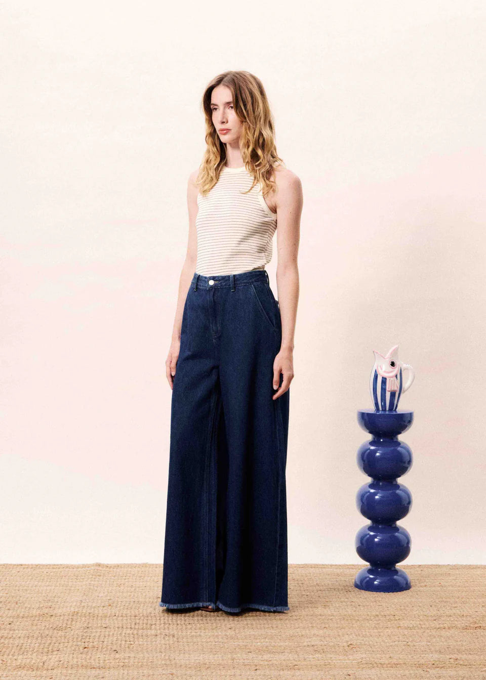 Woman wearing a white sleeveless top and dark blue wide-leg pants standing next to a decorative blue vase.