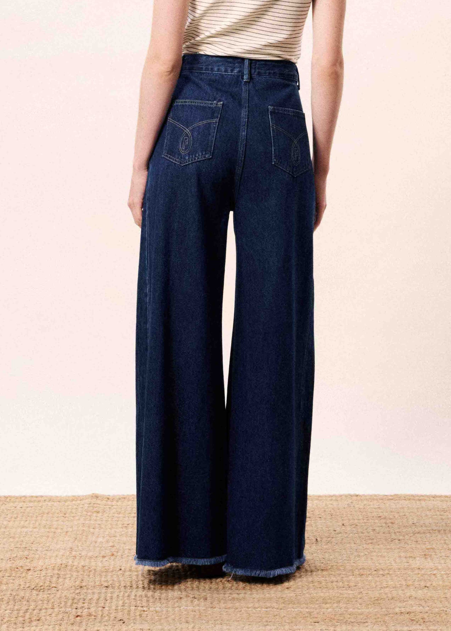 Woman wearing blue jeans standing in front of a white wall with a blue vase on a wooden surface.