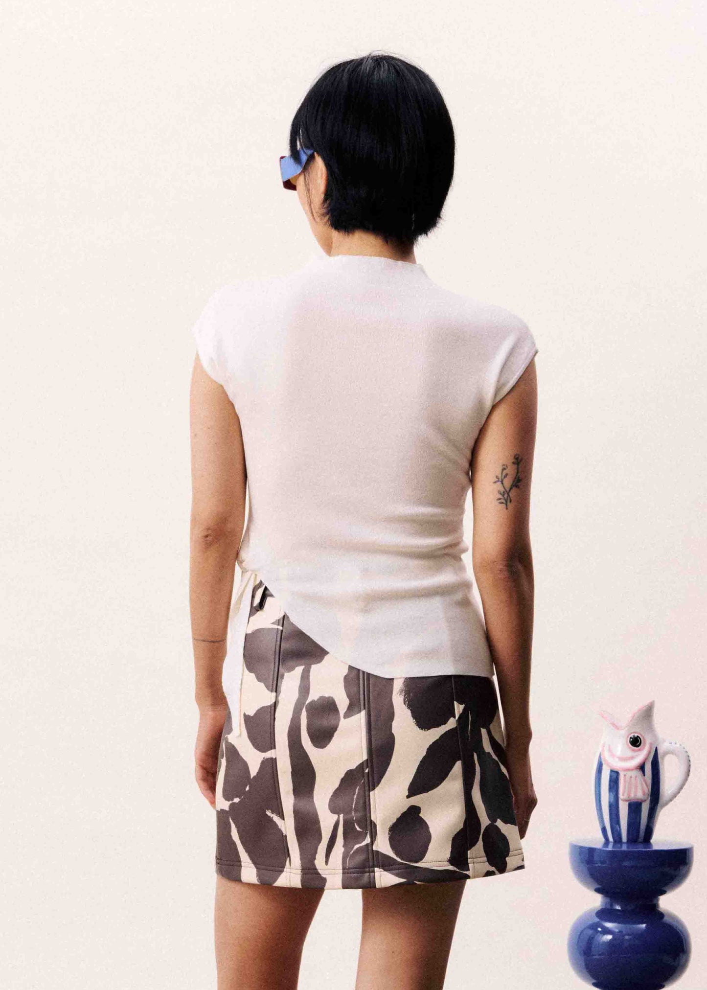Person wearing a white top and patterned skirt standing in front of two small tables.