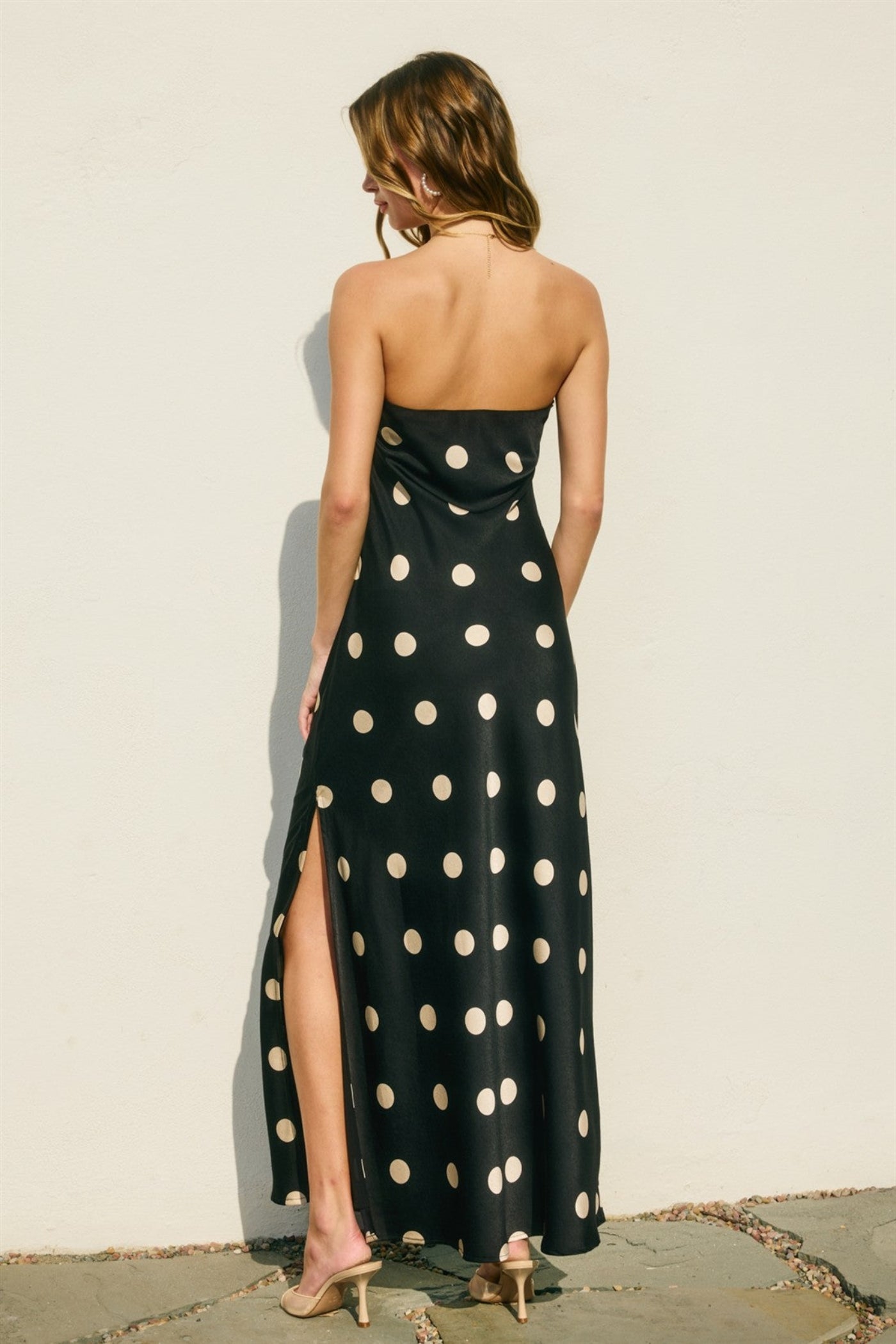 Woman wearing a black strapless dress with white polka dots and a high slit, standing against a light beige wall.