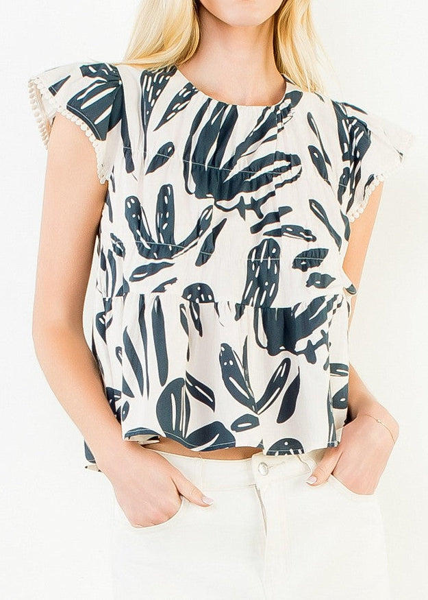 A woman modeling a short-sleeve top with a navy blue botanical leaf pattern on a cream background, paired with a white mini skirt, against a plain white studio background.