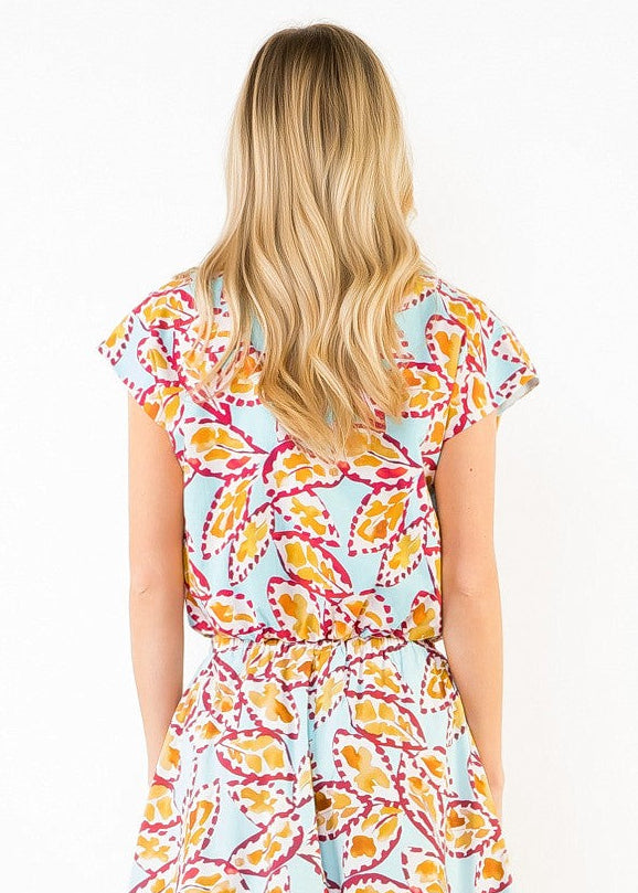 A woman viewed from behind wearing a light blue midi dress with a colorful yellow, orange, and pink floral pattern, paired with beige high-heeled sandals, set against a solid beige background.