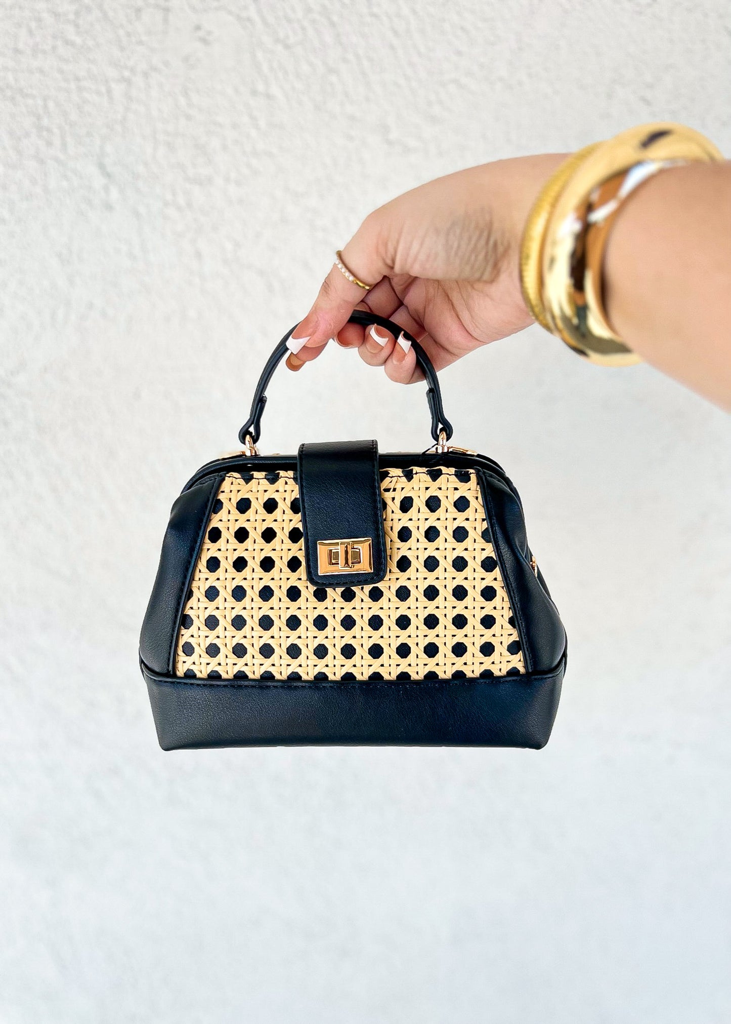 Black and cream woven handbag with black leather trim, metal clasp, and dual handles held by a hand wearing gold bracelets.