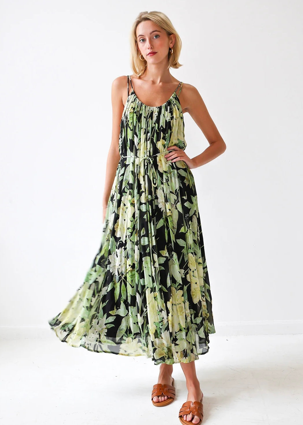Sleeveless maxi dress with green and black floral print, gathered bodice, and midi-length skirt on model wearing tan sandals.