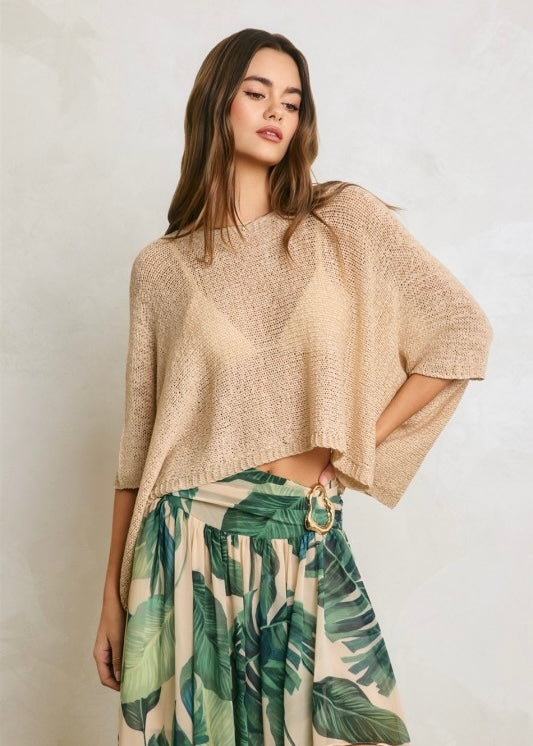 A woman modeling a loose, short-sleeve knit top in a warm beige or oatmeal color, paired with a tropical green leaf print skirt, against a textured light gray wall.