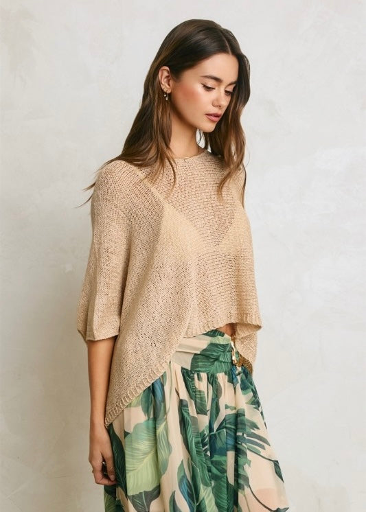 A woman wearing a beige open-knit poncho-style top and a green tropical leaf print skirt, standing against a light textured wall.