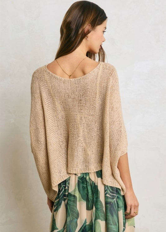 A woman shown from the back wearing a loose, textured knit poncho-style top in a warm beige/tan color, paired with a green tropical leaf print skirt, against a light textured wall.