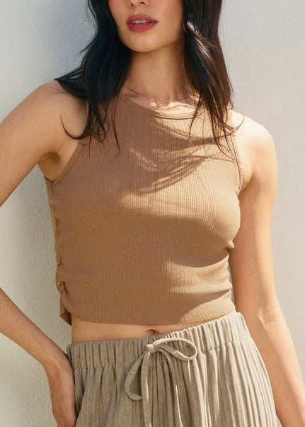 A woman wearing a fitted ribbed sleeveless crop top in a warm taupe-brown color, paired with pleated light beige drawstring bottoms, photographed against a plain light wall with soft natural shadows.