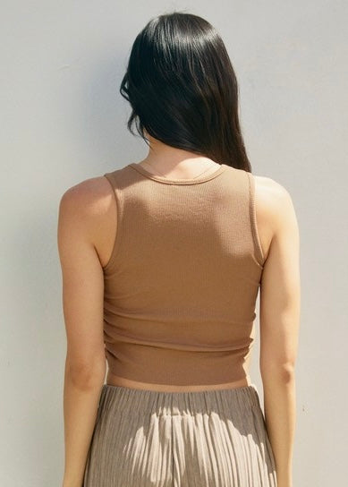 A woman viewed from behind wearing a fitted sleeveless cropped tank top in a warm taupe-brown color, paired with a pleated light taupe skirt, against a plain light gray wall.