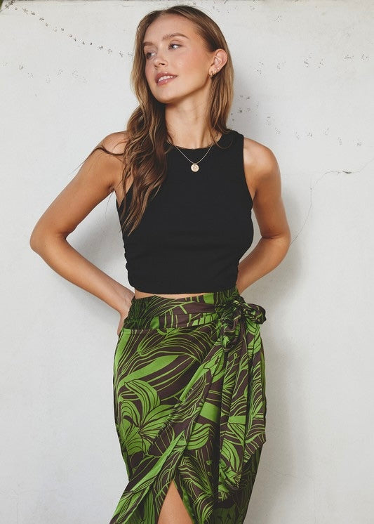 A woman modeling a black sleeveless crop top and a green tropical leaf print wrap skirt against a white textured wall.