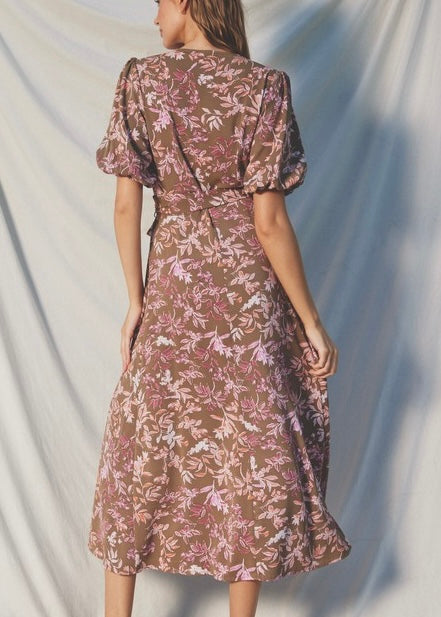 A woman wearing a brown midi dress with a pink and white floral pattern, shown from the back against a draped light gray fabric backdrop.