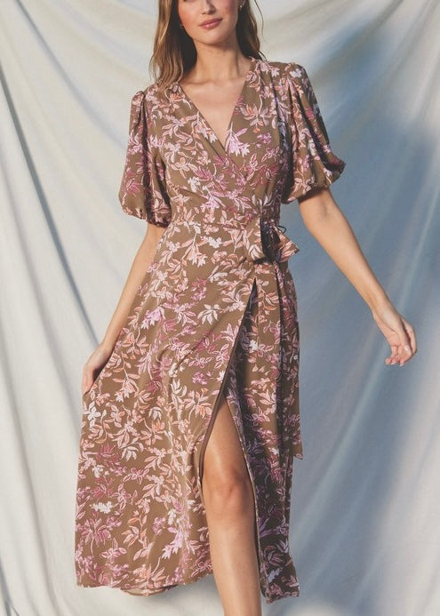 A woman models a brown and pink floral midi dress with puff sleeves, a wrap-style V-neck, and a high side slit, posed against a draped light gray fabric backdrop.