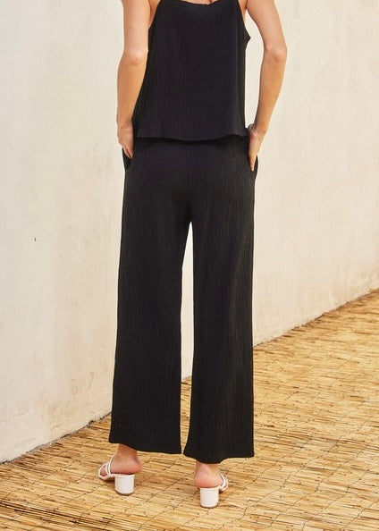 A model from the back wearing a black sleeveless top and matching black wide-leg cropped pants, standing on a woven straw floor against a textured beige wall.