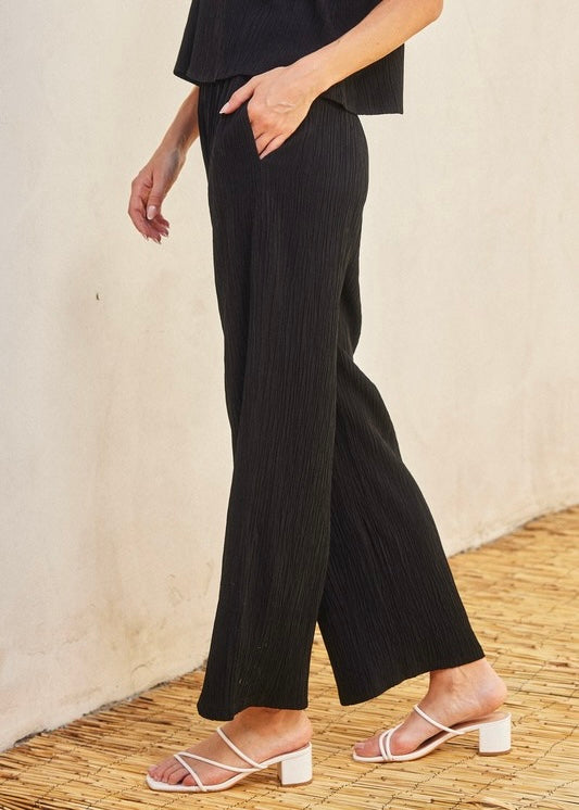 A person wearing black wide-leg pleated trousers with a hand in the pocket, paired with white strappy block-heel sandals, standing against a textured beige wall and woven floor.