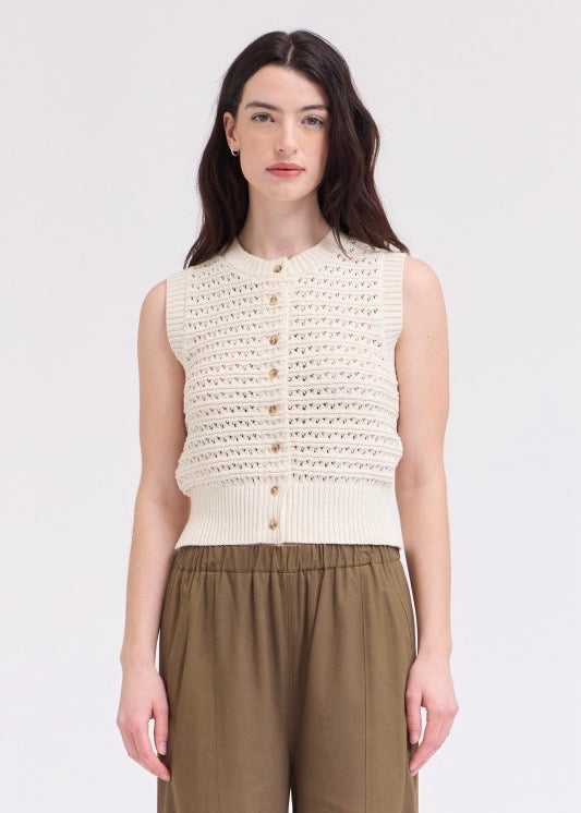 A woman modeling a sleeveless cream-colored knit vest with a button-down front, paired with olive green trousers, against a plain light gray background.
