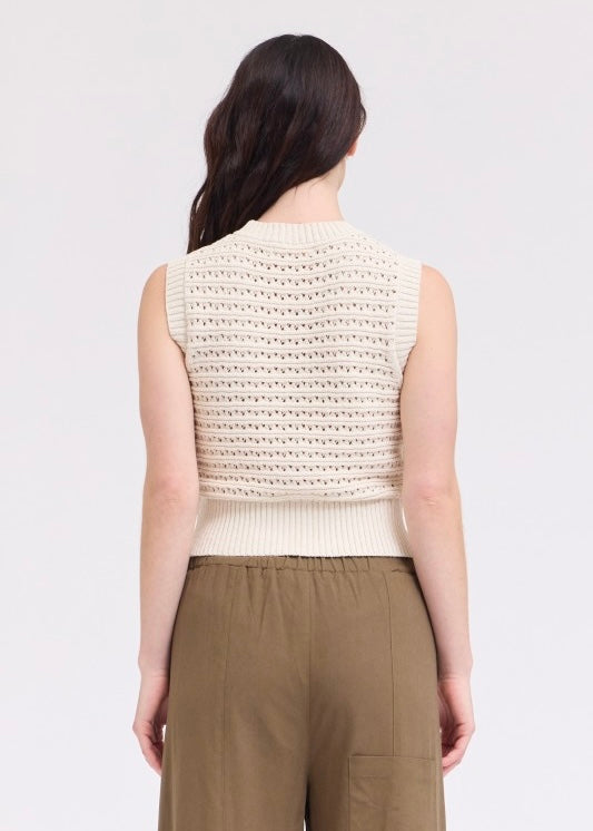 A woman shown from the back wearing a sleeveless cream knit vest with a textured openwork pattern and olive green pants, against a plain light gray studio background.