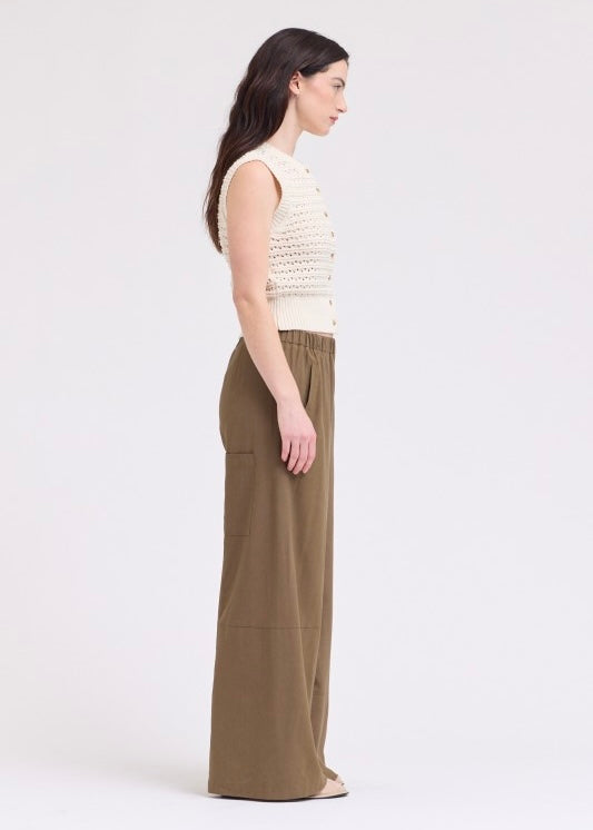 A woman shown in side profile wearing a sleeveless cream knit top and olive green wide-leg trousers against a plain light gray studio background.