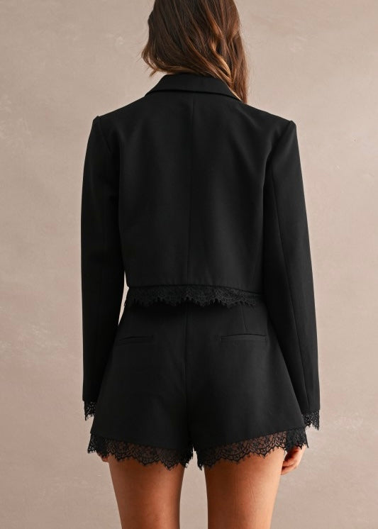 A model shown from the back wearing a black cropped blazer and matching black shorts with delicate black lace trim along the hem and cuffs, set against a neutral textured beige background.