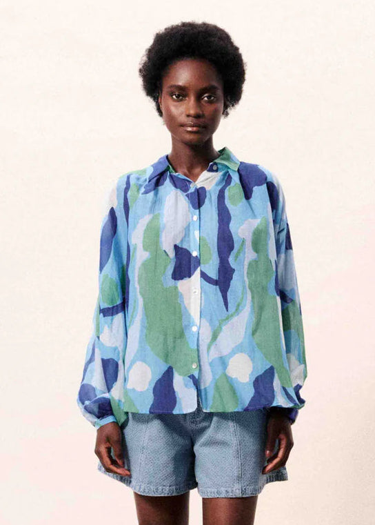 A model wearing a long-sleeve button-up shirt with a blue, green, and white abstract pattern, paired with light-wash denim shorts, against a plain light beige background.