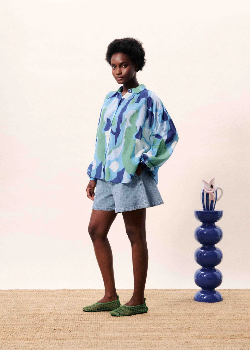 Person wearing a colorful shirt and shorts standing next to a blue vase on a wooden floor.