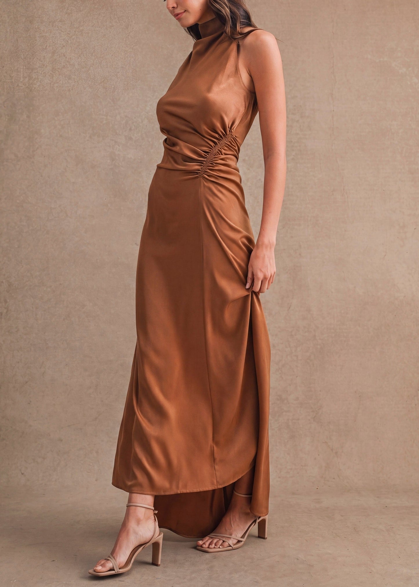 A woman models a sleeveless, high-neck, floor-length satin dress in a warm camel brown color, standing against a textured beige studio backdrop.