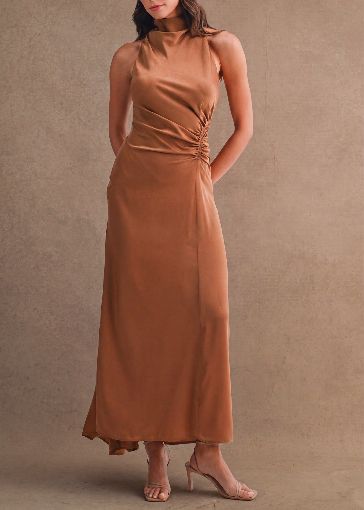 A woman models a sleeveless, high-neck, floor-length brown dress with gathered detailing at the waist, standing against a textured brown studio backdrop.