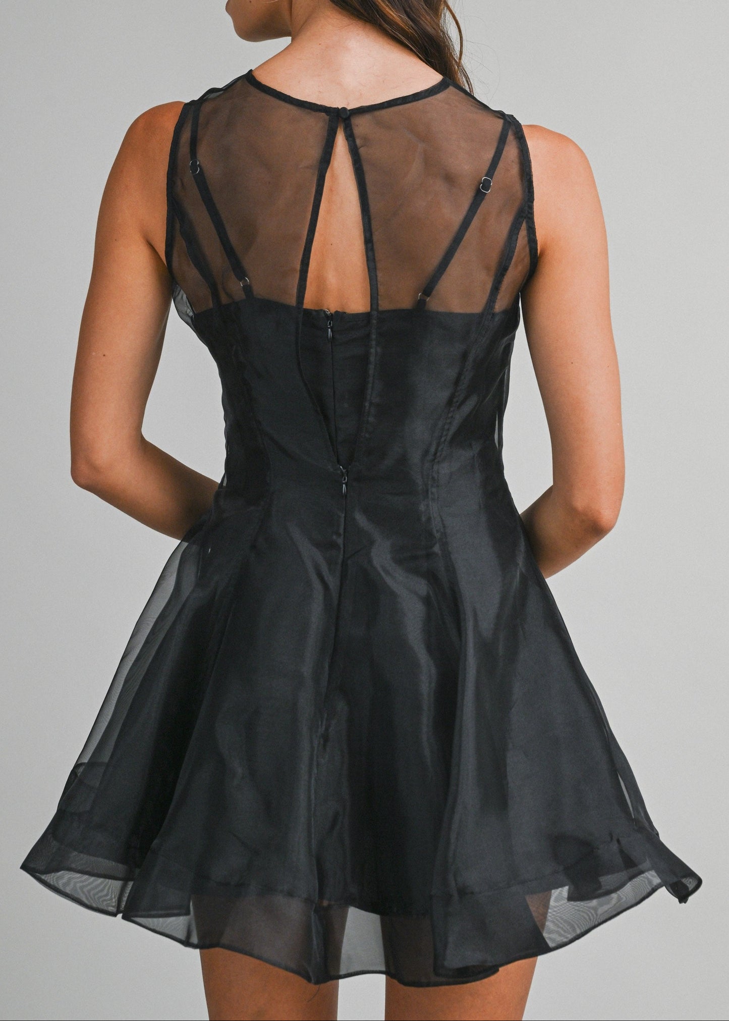 A woman shown from the back wearing a black sleeveless dress with sheer mesh back panels and thin crisscross straps, photographed against a plain light gray studio background.