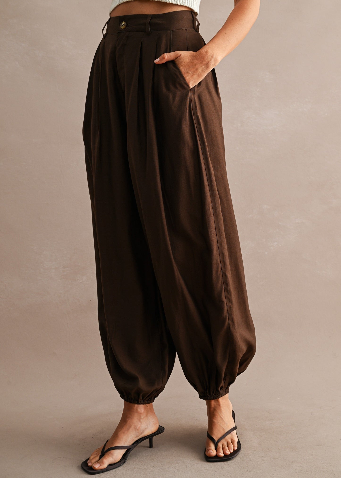 A model wearing high-waisted, voluminous brown harem-style pants with side pockets, paired with a white ribbed crop top and black thong sandals, set against a neutral textured studio backdrop.