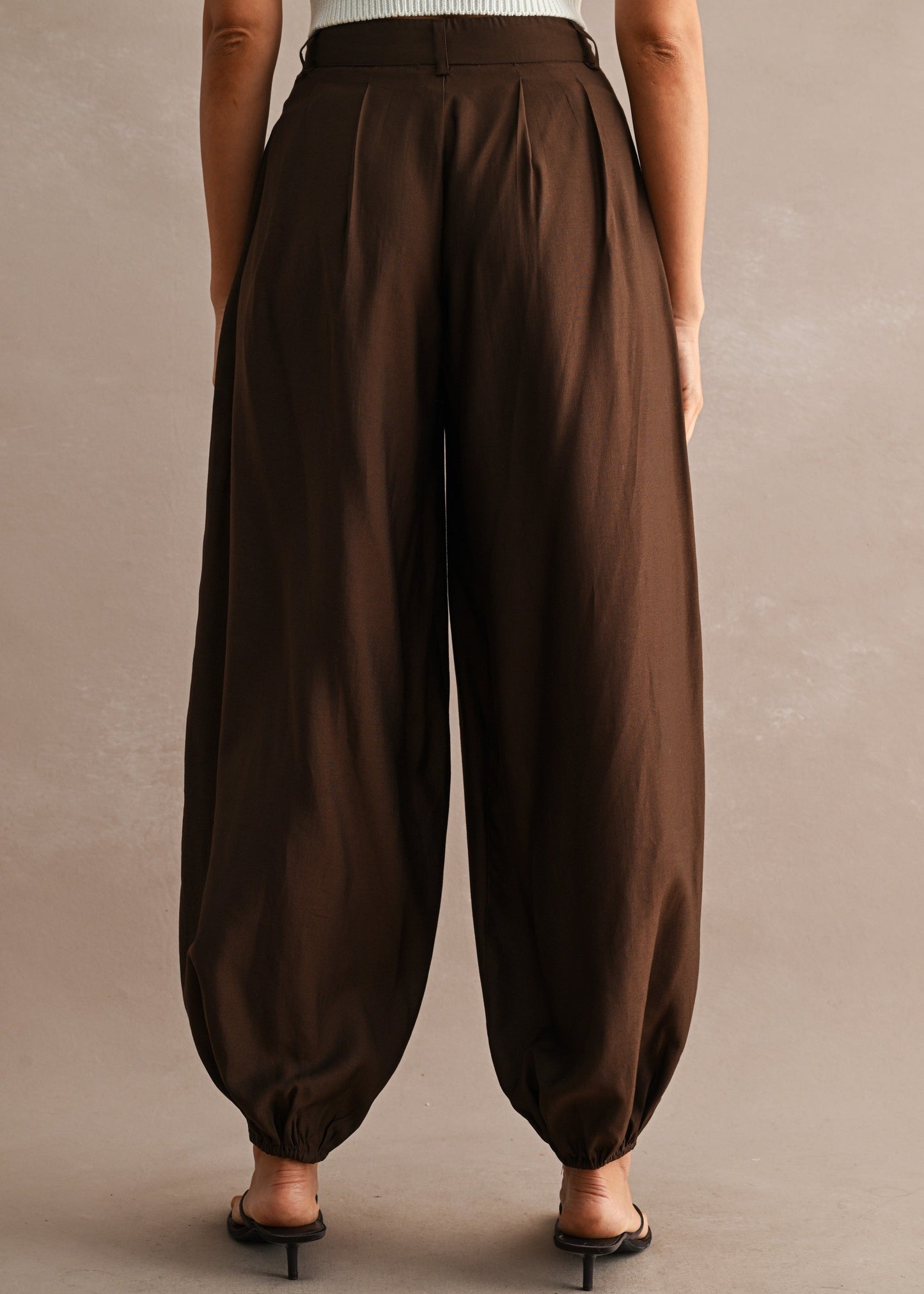 A person shown from the back wearing loose, high-waisted brown harem-style pants with gathered cuffs, paired with a white ribbed top and black heeled sandals, against a plain warm beige studio background.