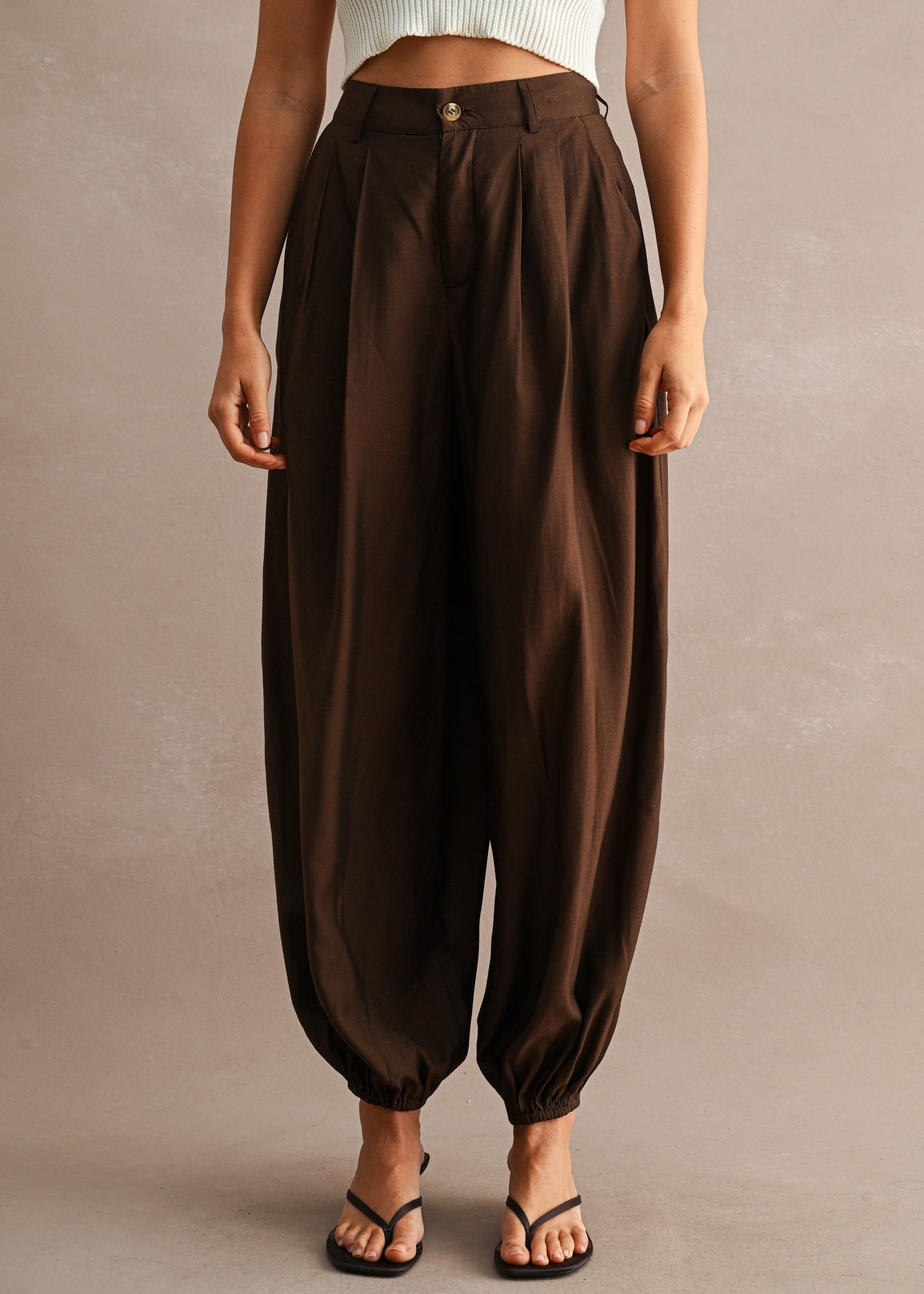 A model wearing high-waisted, loose-fitting brown harem-style pants with gathered legs and elastic ankle cuffs, paired with a white ribbed crop top and black thong sandals, against a textured beige studio backdrop.