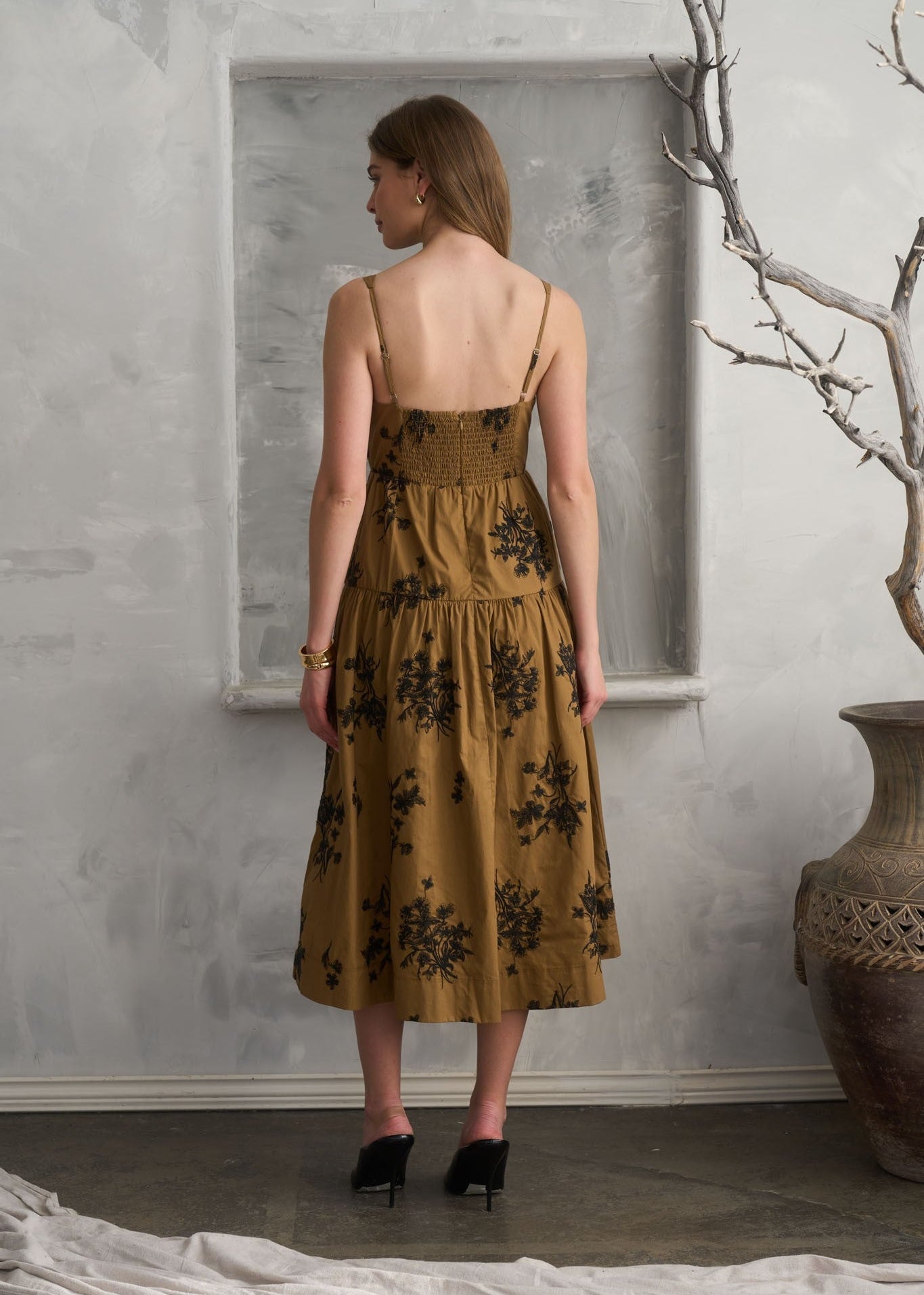 A woman stands with her back to the camera wearing a sleeveless midi dress in a mustard yellow color with a black floral pattern. She is standing in front of a textured gray wall with a large recessed panel, next to a large ceramic vase with a bare branch.