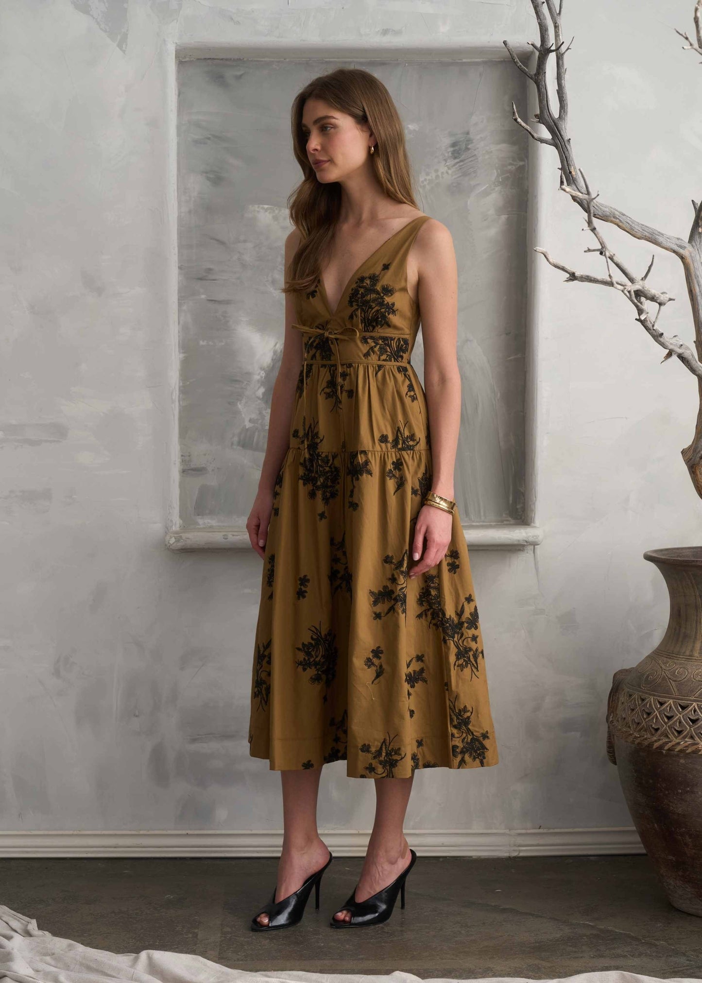 A woman models a sleeveless midi dress with a deep V-neckline, a fitted waist, and a flowing skirt. The dress is a warm mustard yellow color with a black floral pattern.