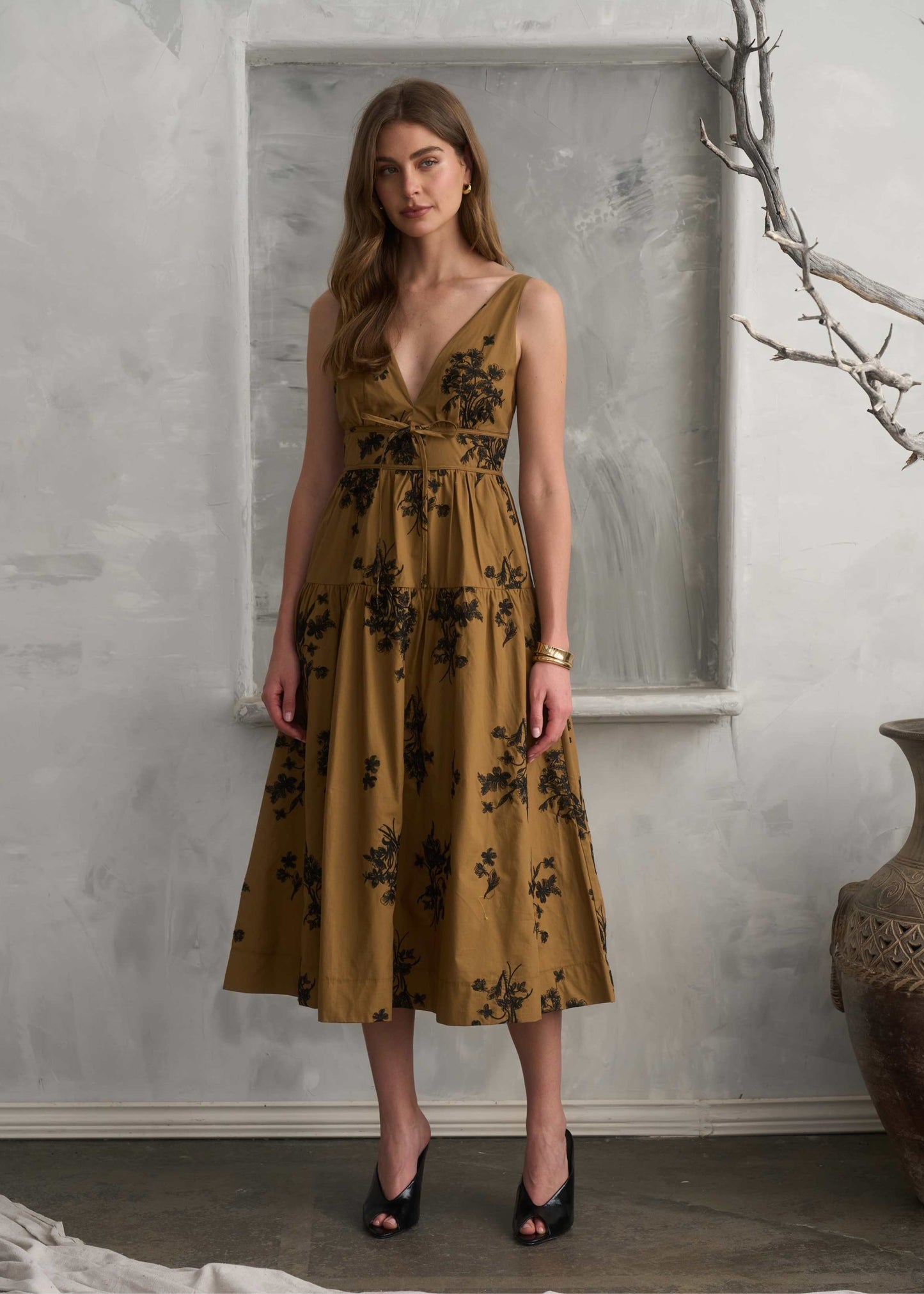A woman models a sleeveless, V-neck midi dress in a mustard yellow color with a black floral pattern, standing in front of a textured gray wall.