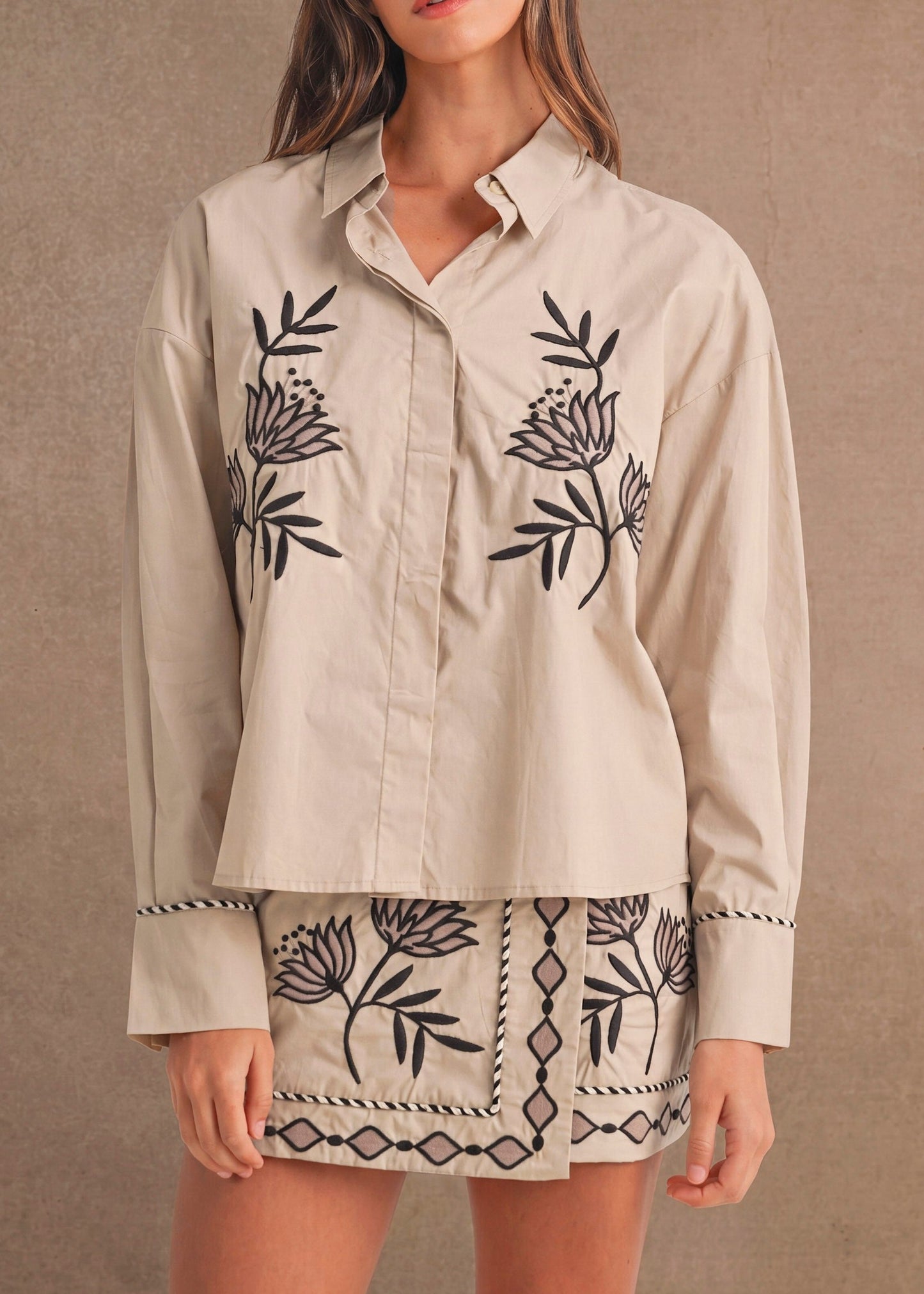 A woman modeling a beige long-sleeve button-up shirt with black floral embroidery and a matching embroidered mini skirt, set against a textured beige studio backdrop.
