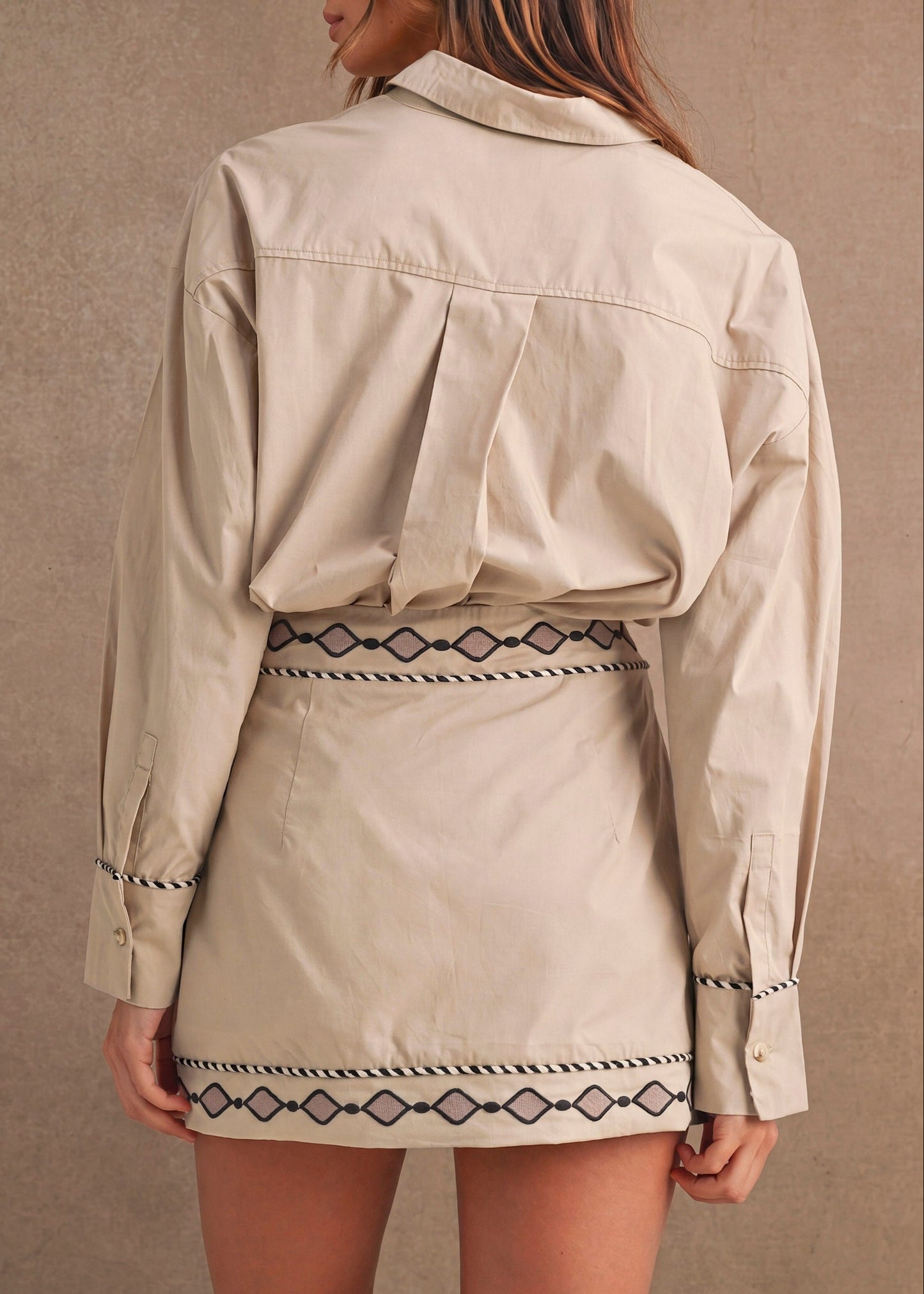 A woman shown from the back wearing a light beige cropped utility jacket and matching mini skirt with black geometric embroidered trim, set against a textured beige wall.