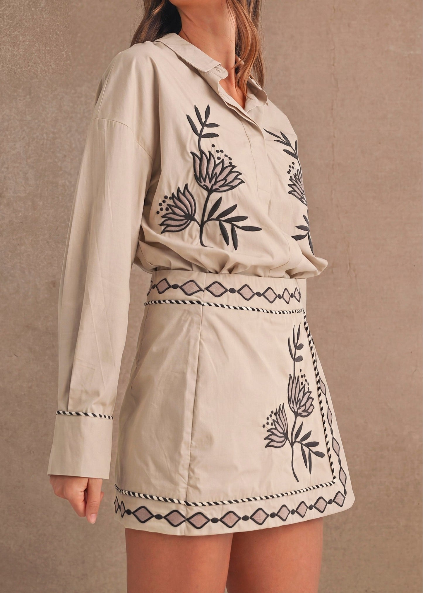 A woman modeling a beige long-sleeve shirt and matching mini skirt set with black embroidered botanical and geometric patterns, set against a textured beige wall.