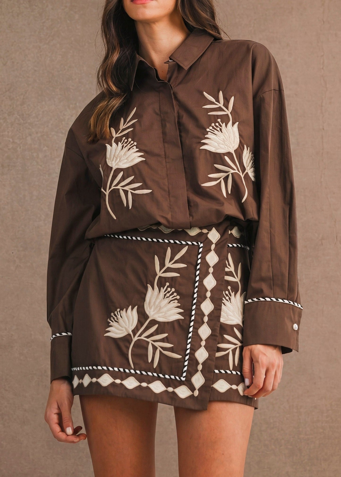 A woman modeling a brown button-up shirt and matching mini skirt set with white floral and geometric embroidery, set against a textured brown studio backdrop.