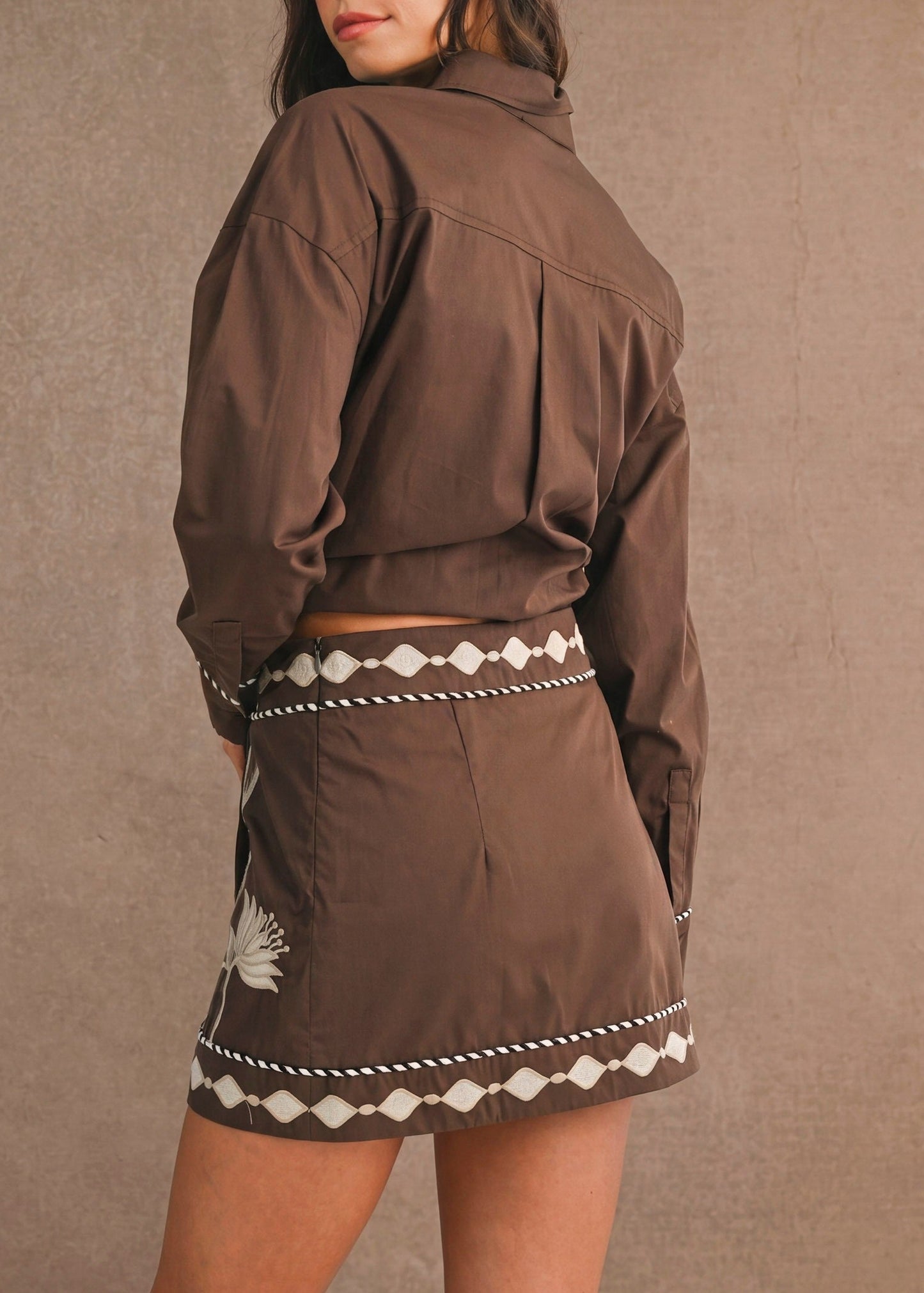 A woman wearing a brown cropped jacket and matching mini skirt with white geometric trim, photographed against a textured brown backdrop.