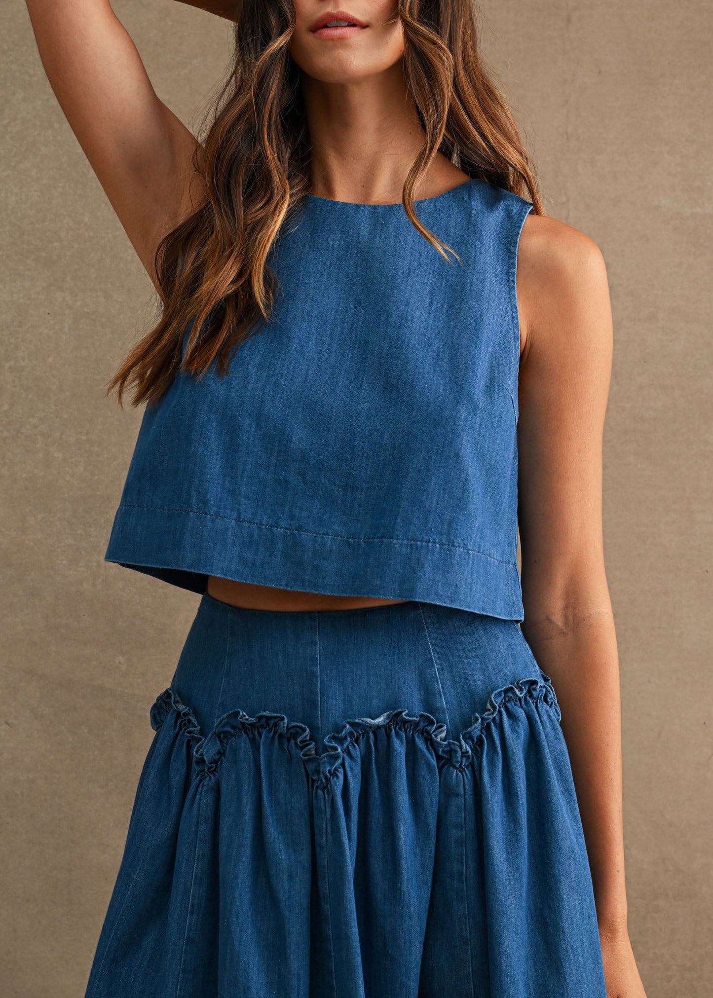 A woman modeling a sleeveless  denim blue cropped top and matching blue high-waisted skirt against a textured beige studio backdrop.