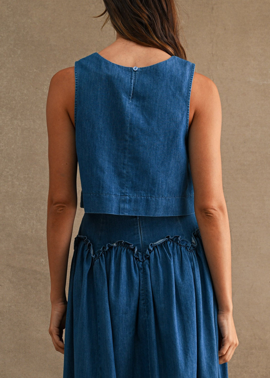 A woman viewed from the back wearing a sleeveless denim top with a fitted bodice and gathered skirt, standing against a plain warm beige studio backdrop.