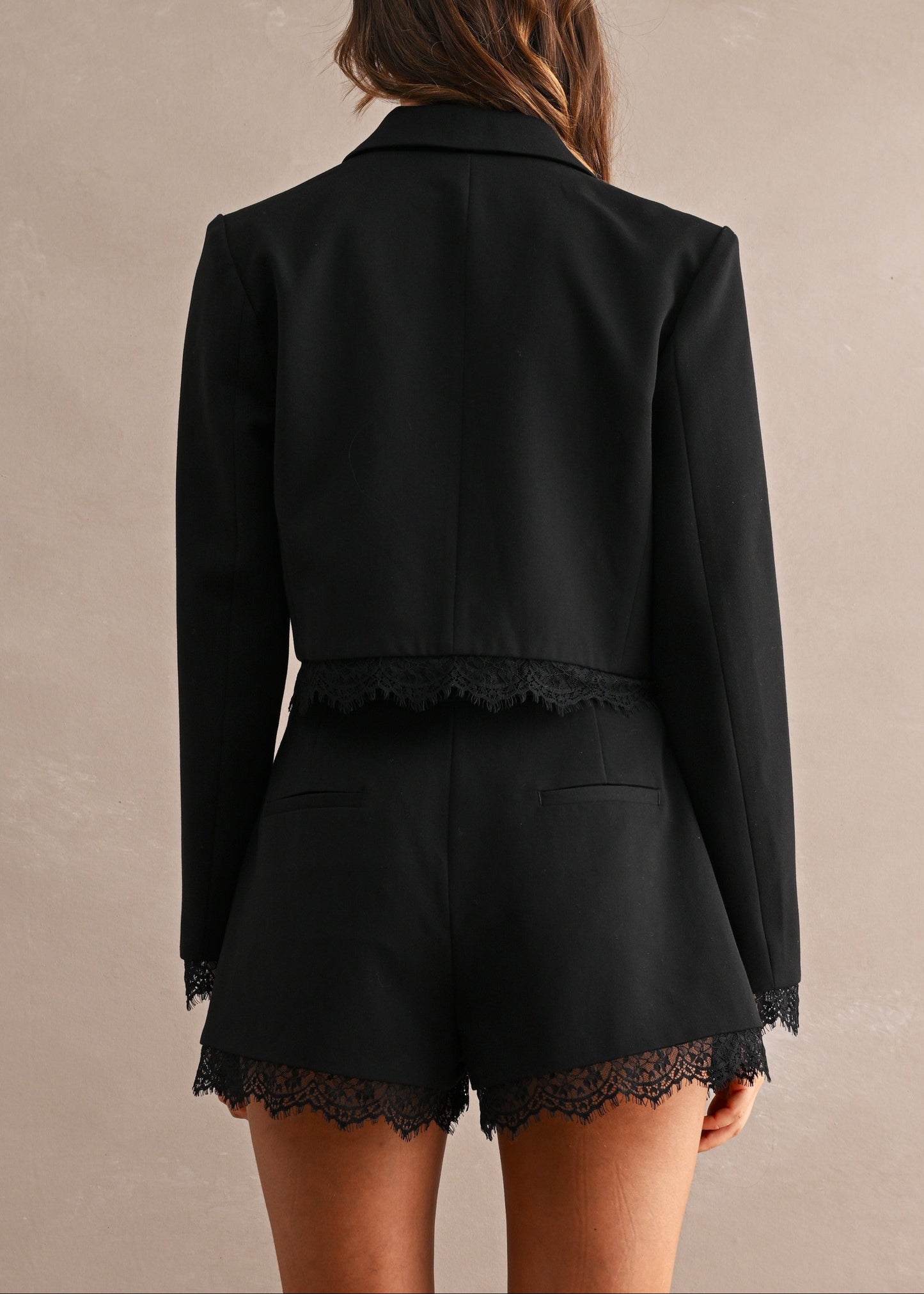 A woman shown from the back wearing a black cropped blazer and matching black shorts with lace trim, against a plain warm beige studio background.