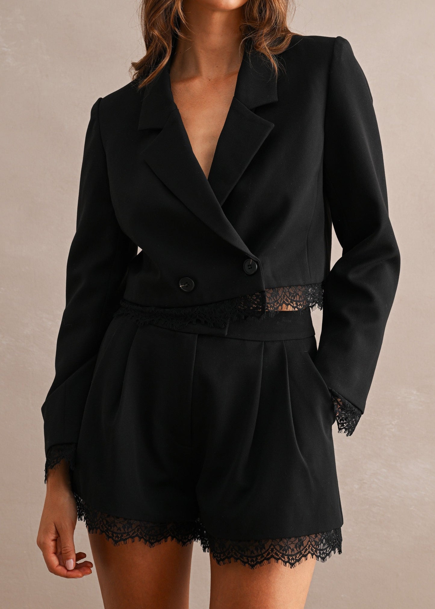 A woman modeling a black double-breasted blazer and matching shorts set with lace trim, photographed against a neutral textured beige studio backdrop.