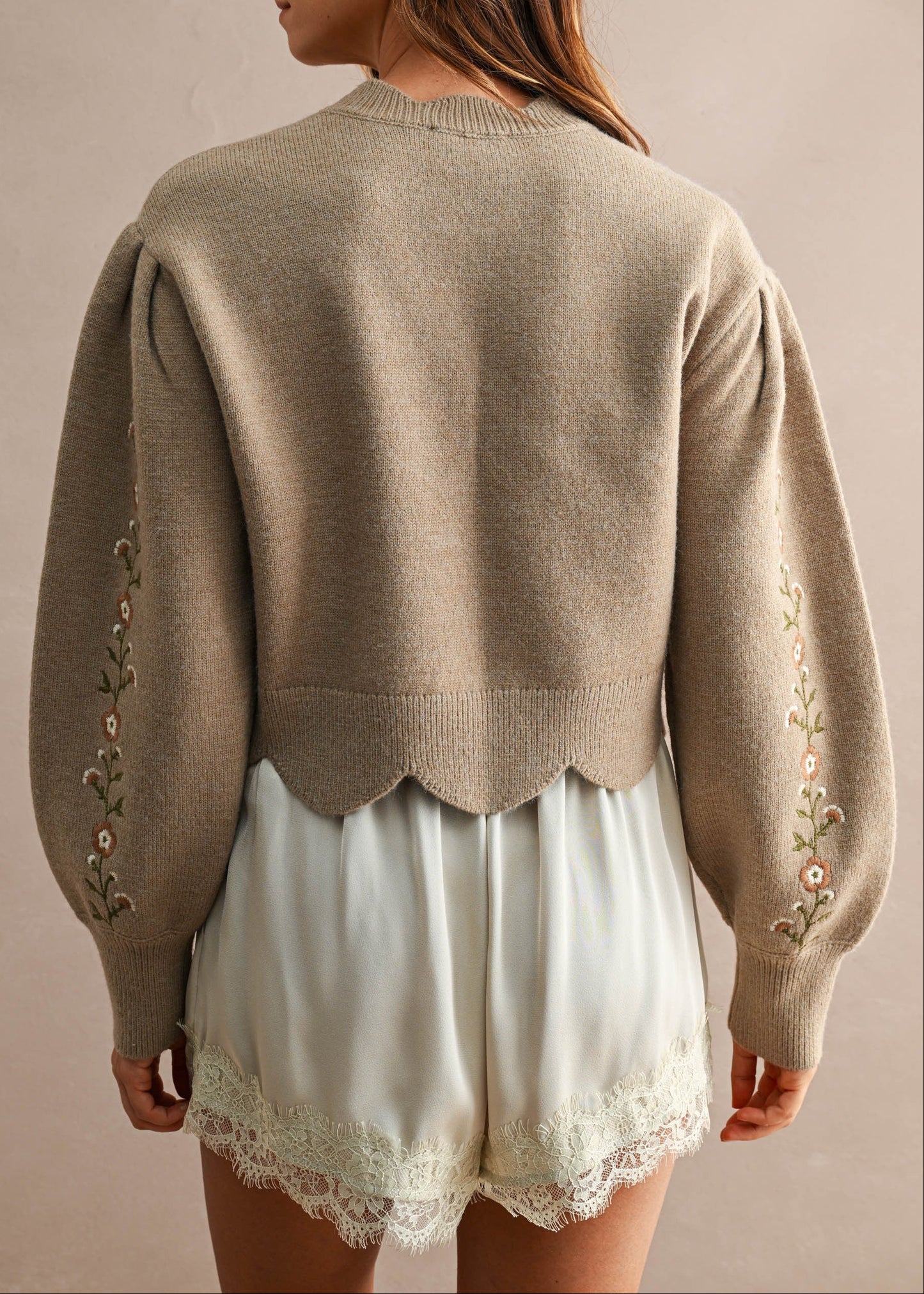 A woman shown from the back wearing a taupe knit sweater with decorative floral embroidery down the sleeves and a scalloped hem, paired with white lace-trimmed shorts against a neutral beige background.